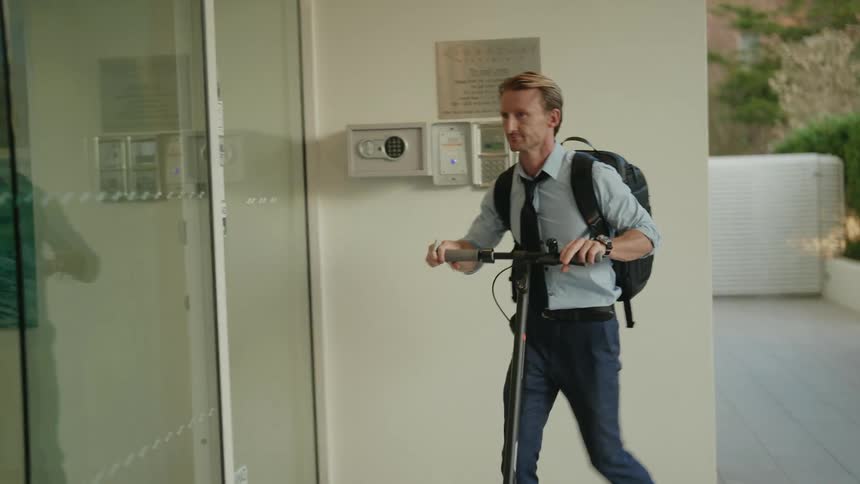 A man is walking with his scooter to a building and he pushes a button before he goes inside