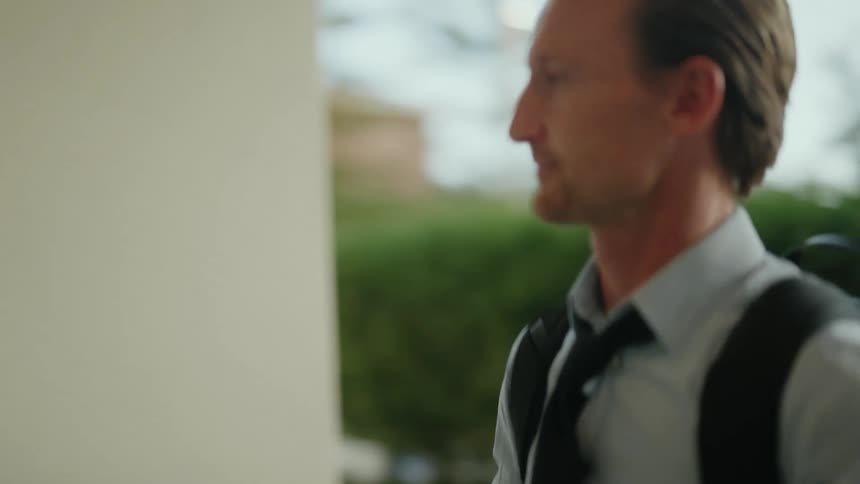 A man wearing a tie and a backpack is walking his scooter to a building where he pushes a button on a panel on the wall