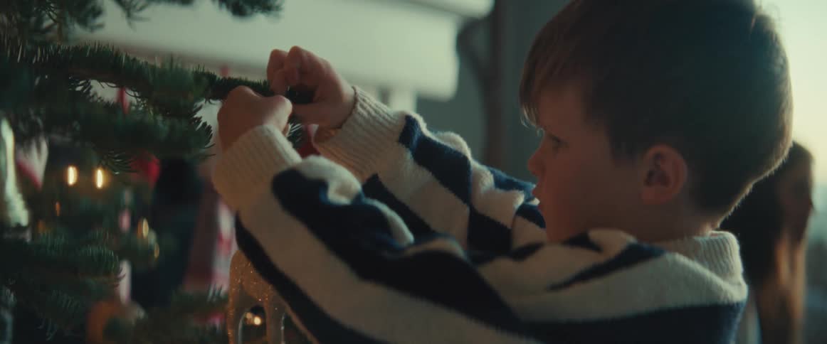 A little boy is hanging a reindeer decoration on a christmas tree as his mother walks in the distance