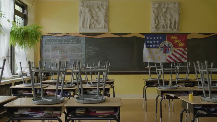 Stools are on top of the desks in an empty classroom with plants hanging near the windows and a flag is on the blackboard