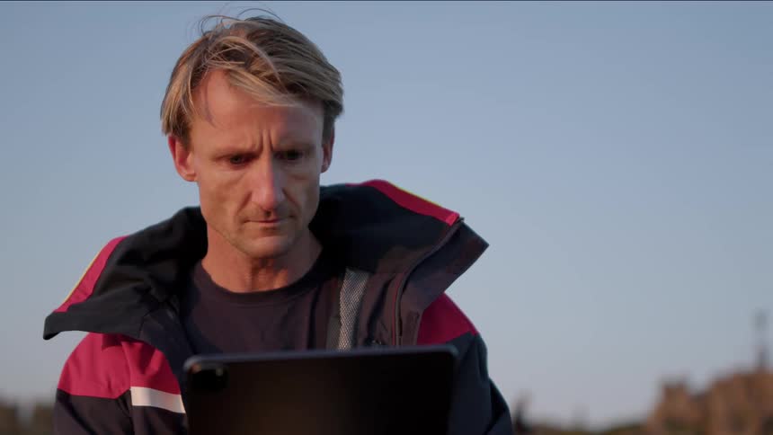 A man is standing outside holding a tablet and he looks at his watch