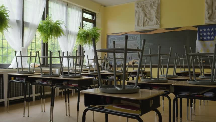 Stools are on top of the desks in an empty classroom with plants hanging near the windows