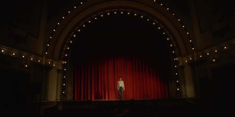 A man walks out onto a stage in a dim theatre