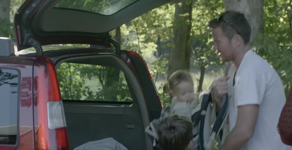 A man with several children lifts a baby in a carrier onto his back