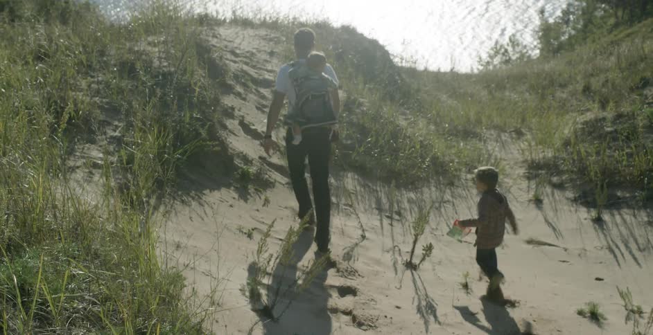 A man carries a baby and his back and is trailed by a boy as he walks