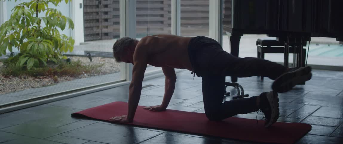 Topless muscular man works out on red yoga mat next to grand piano in room