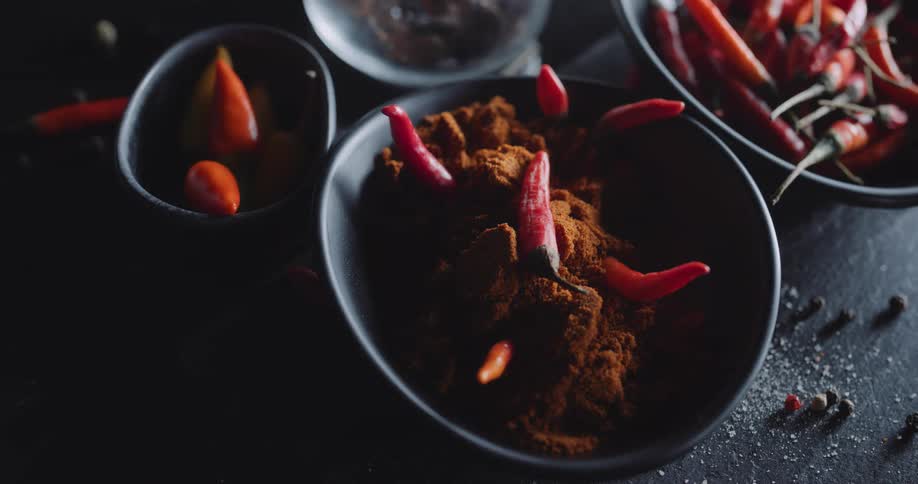 Red chili peppers sit alone or with chili powder in black bowls on black surface scattered with peppercorns and salt