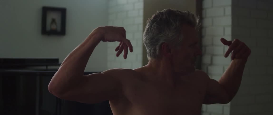 A bare chested man is standing in a room looking from side to side as he raises his arms in the air