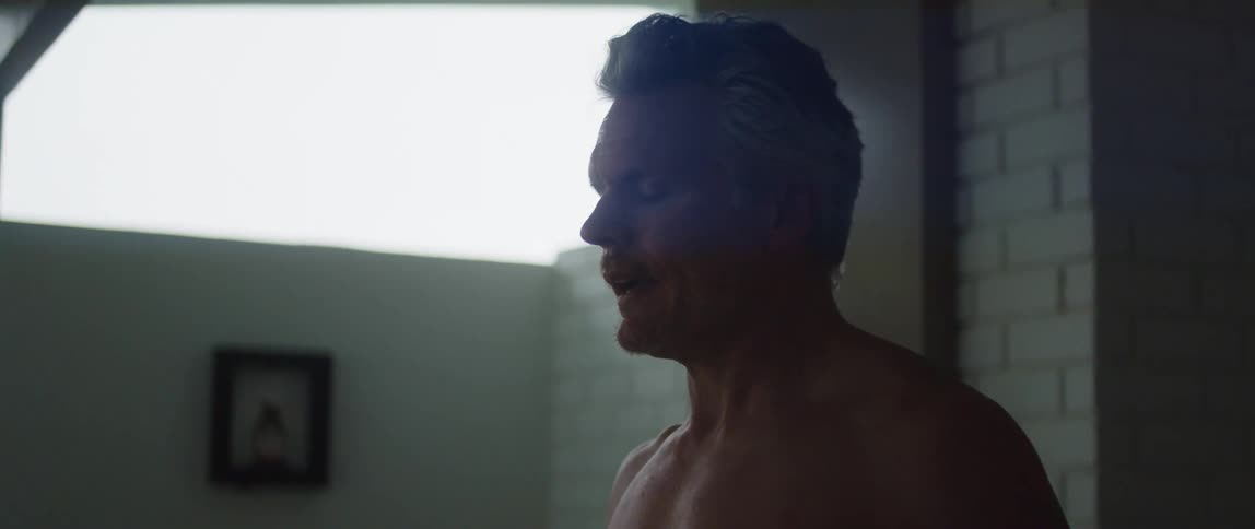 A bare chested man is standing in a room breathing deeply as he raises his arms in the air