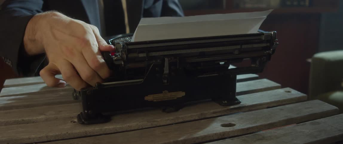 Man with black jacket sits at desk using typewriter in room