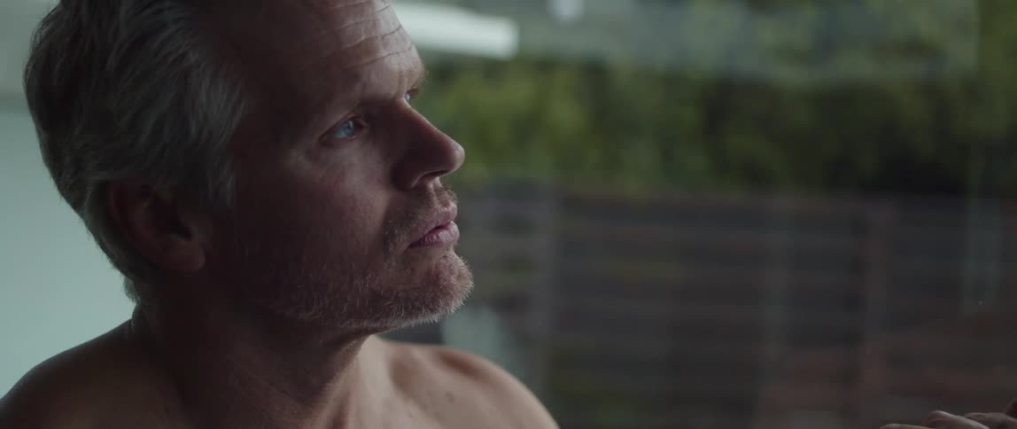 A bare chested man is standing in a room near a window as he takes a deep breath