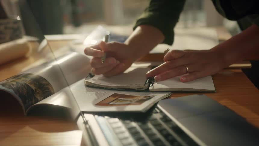 A woman is standing over a table writing in a notebook near her laptop computer