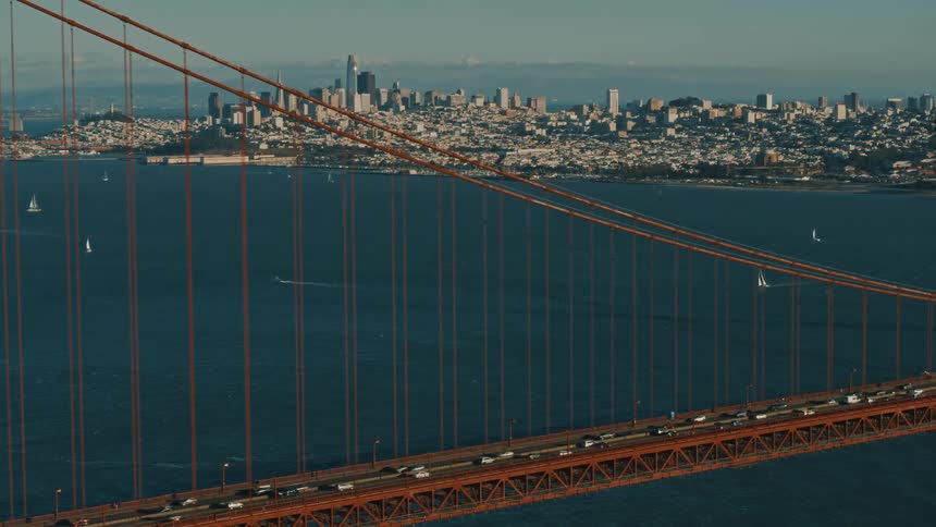 An aerial view of vehicles traveling across the Golden Gate Bridge with sailboats in the water and the San Francisco skyline in the distance