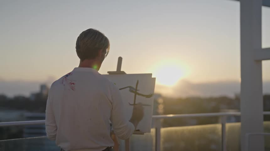 Male painter stands on balcony painting scene at sunset