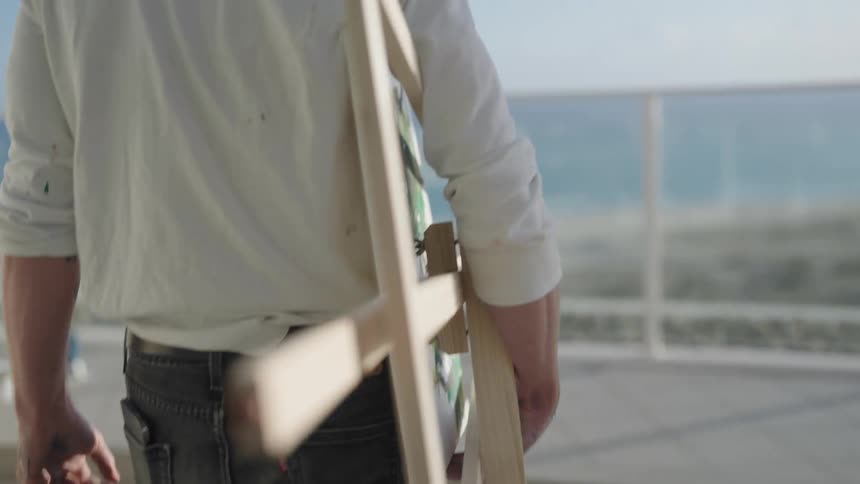 A man at home carries an easel on to his balcony off the water to paint