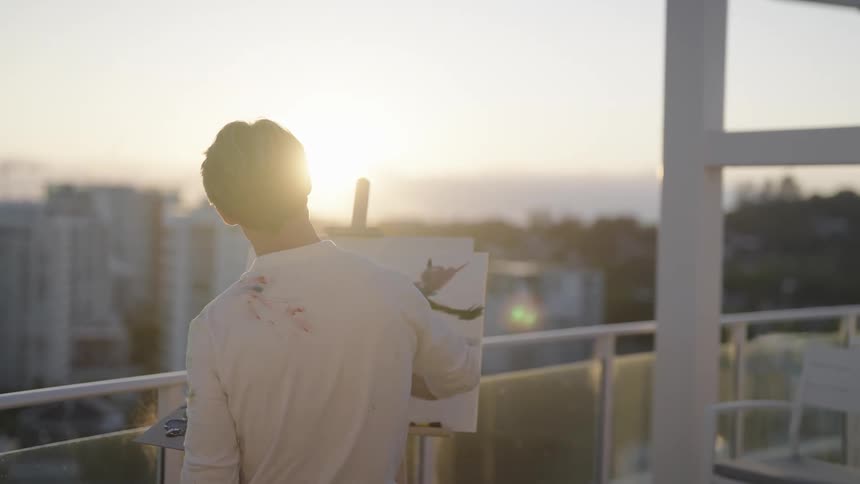 Man with white shirt paints on a canvas on balcony at sunset