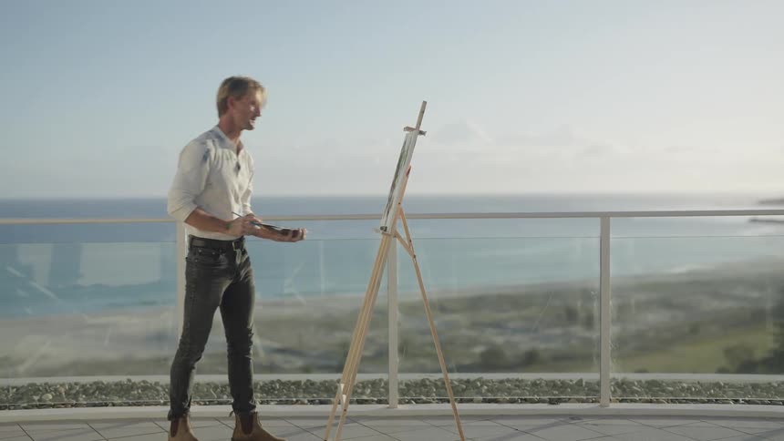 A man stands on the shore and paints on a canvas