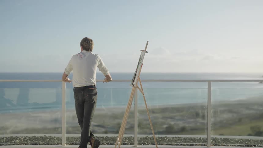 Man studies painting on easel before leaning against banister to look at ocean view