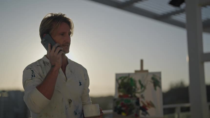 Blonde man in white and paint stains shirt talks on smartphone and drinks from cup in front of canvas on balcony as the sun shines brightly