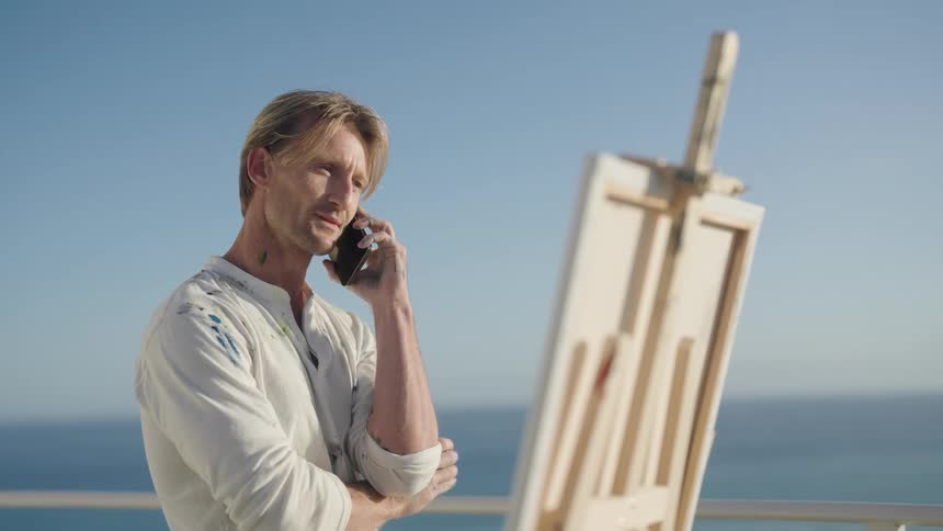 Paint splattered artist holds cup and talks on smartphone next to painting on easel with ocean in the background