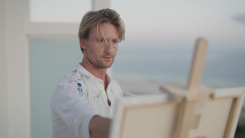 A man steps back to look at his painting on an easel