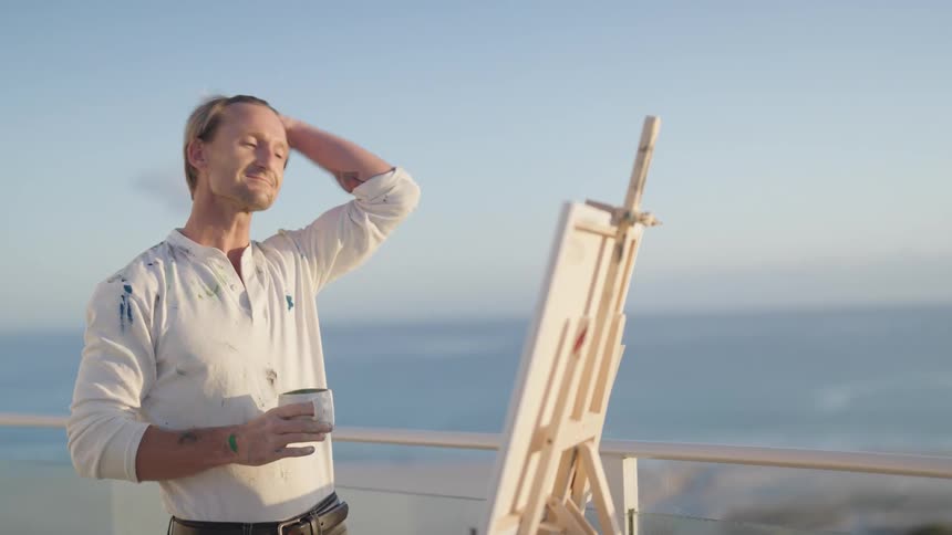 A man stands on his balcony on the shore and takes a drink of coffee and looks at his painting on an easel