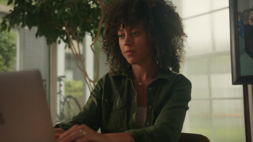Woman with curly hair denim shirt types on laptop and rubs her tired hands sitting at desk