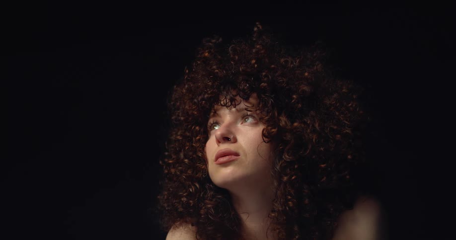 Light shines on the face of a woman with long dark curly hair as she looks up from a dark room