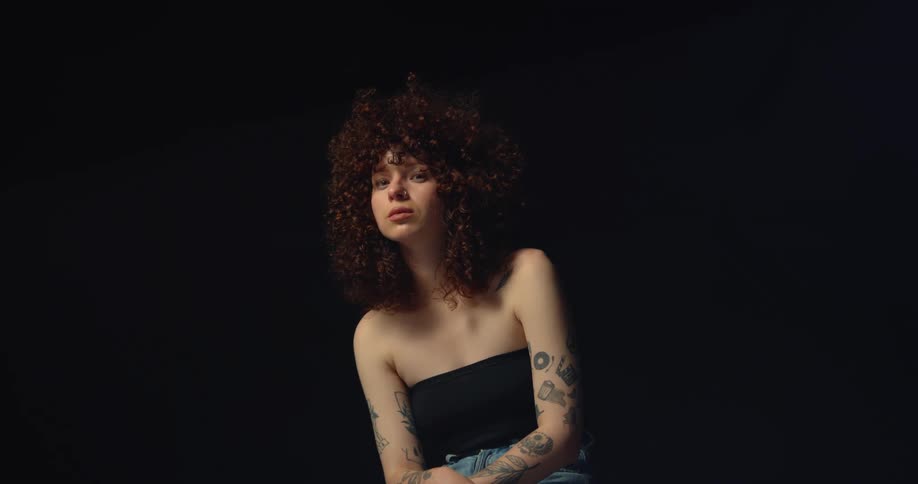 A woman with incredibly curly hair and tattoos poses while sitting for the camera