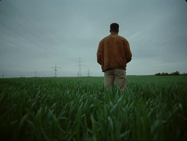 Man in brown jacket and pants stands in green grass on a cloudy day