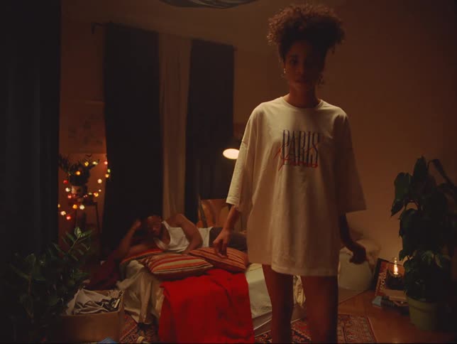 A man lies on a bed reading while a woman backs up to look at herself in a mirror