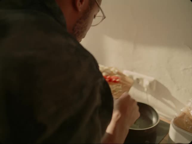 Man with beard glasses and green shirt puts chopped onions garlic and tomatoes in pan in the kitchen
