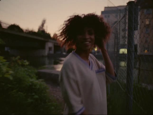 A teenage girl is walking near a chain link fence toward apartment buildings and she turns around