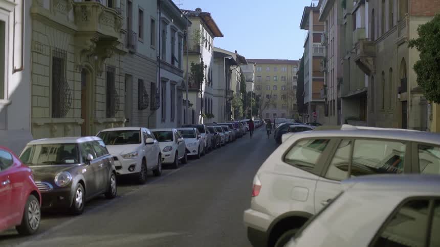 Two people ride bikes down a road lined with cars