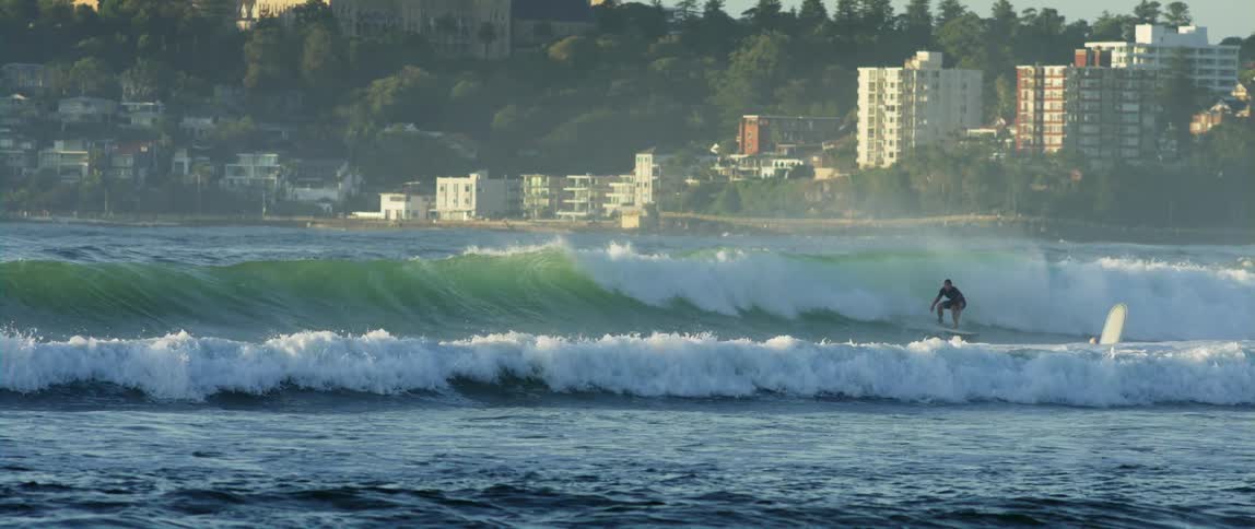 Surfer rides a wave with buildings and trees in the background