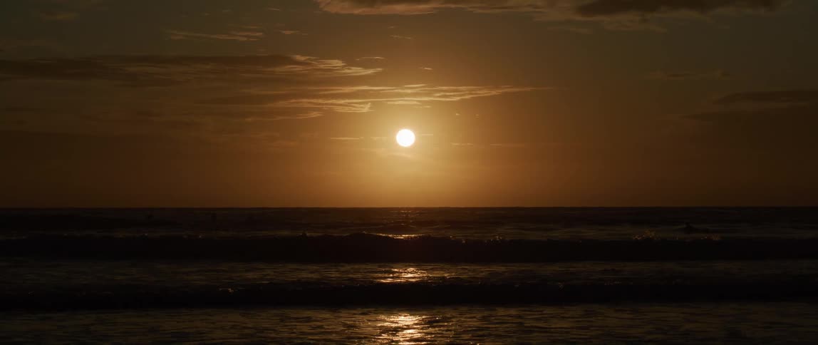 Waves ripple and reflect light as they roll into shore and the sun is setting in an orange sky