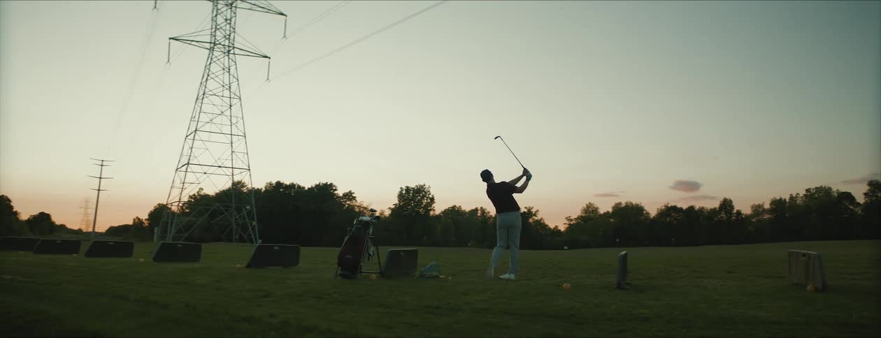 Man swings golf club at driving range under power lines at dusk