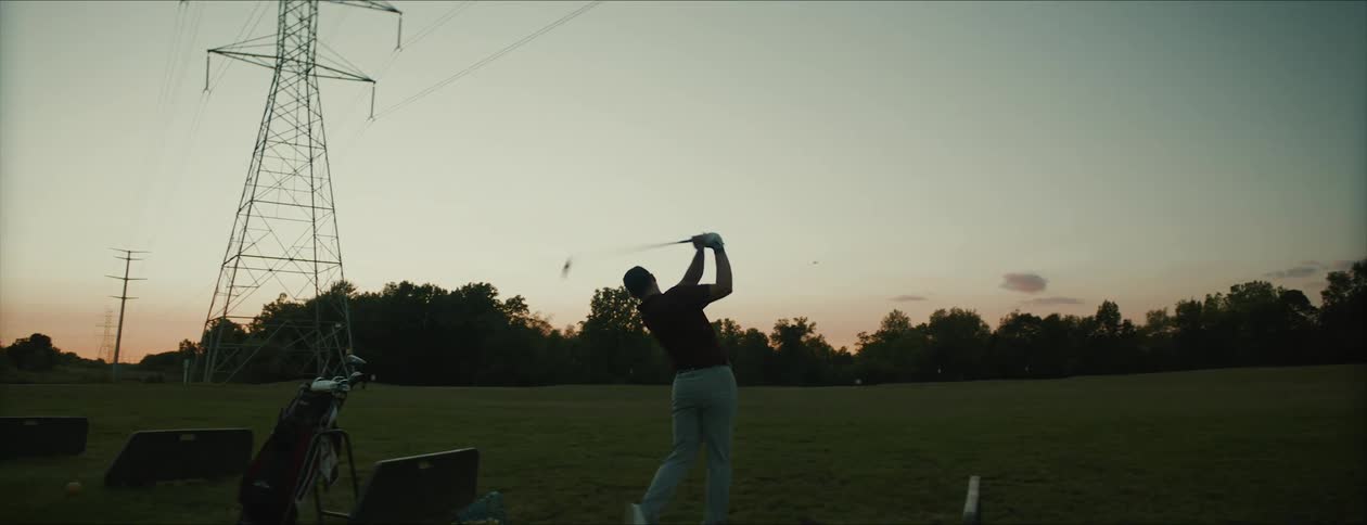 Male golfer drives ball across field under power lines under a red sky