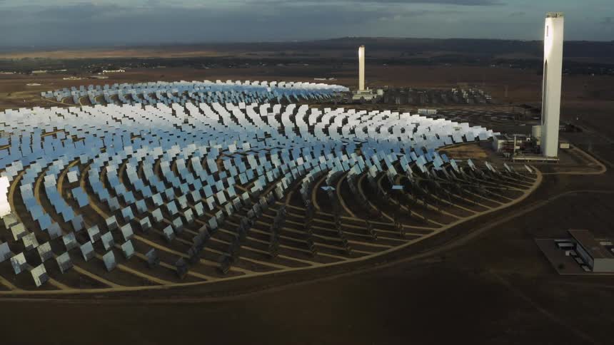 Tall solar collector towers gather sun rays from a circular field of solar panels lined up in neat rows below