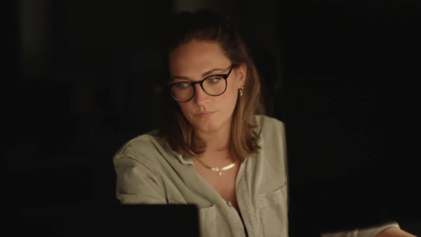 A woman is sitting at a desk working on two computers and turning to look at the other monitor
