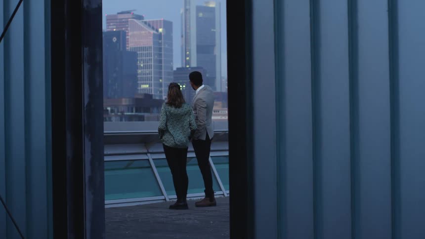 Couple stands on roof as man gestures toward skyscrapers