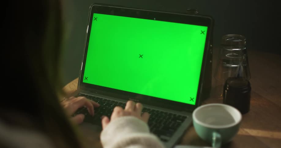 A woman is sitting at a table using a laptop computer with a green screen