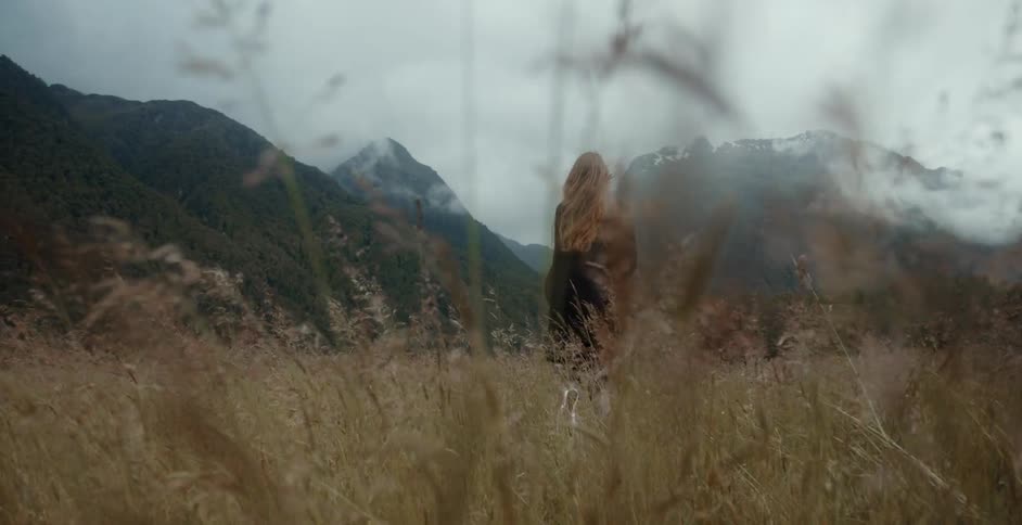 A long haired female walking in a field near mountains