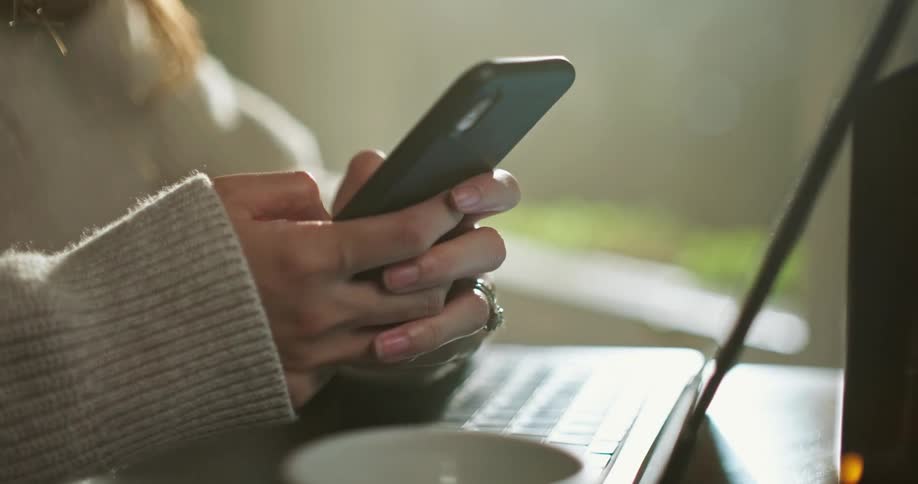 A woman is sitting at a table with a laptop computer texting on her phone