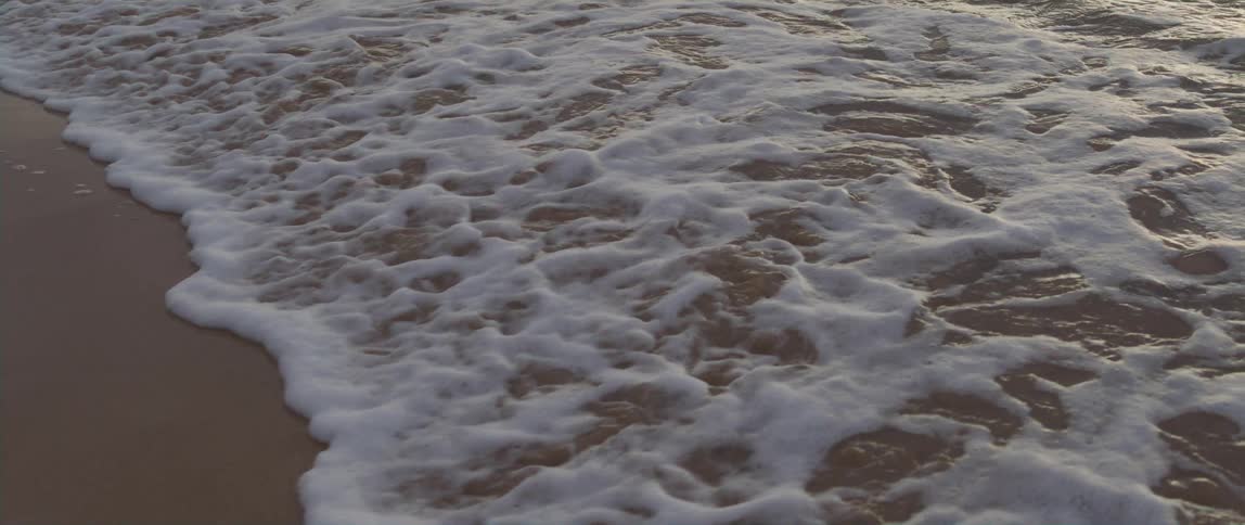 Slow motion footage of small ocean waves washing over the wet sand at evening