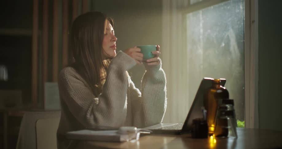 Young woman with laptop drinking coffee at sunrise in cafe