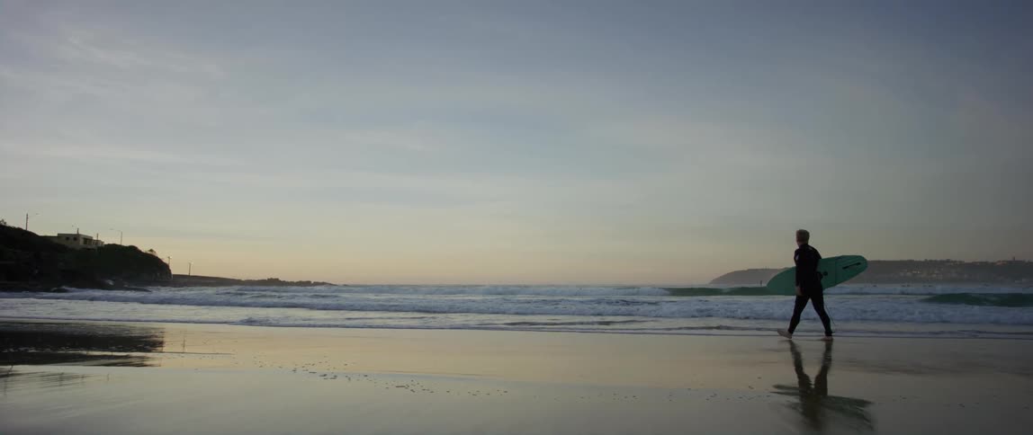 A man is walking across the beach carrying his surfboard into the water