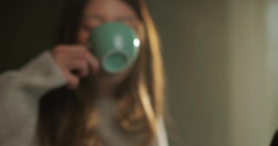 A woman typing on a laptop at a desk stops to drink from a coffee mug and gaze at the nearby window