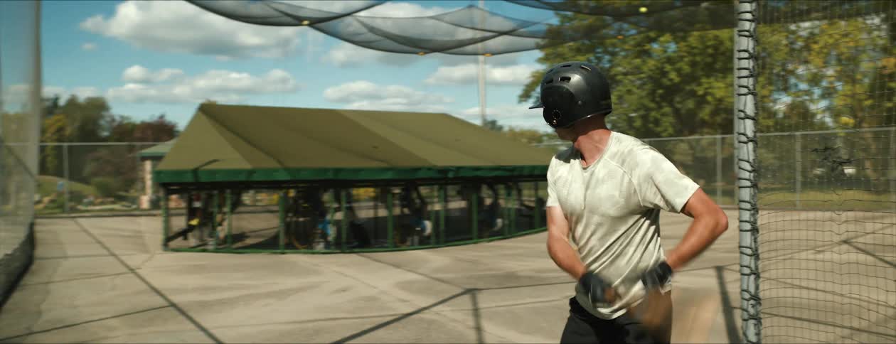Batters standing in batting cages swing at balls pitched by machines as another person watches