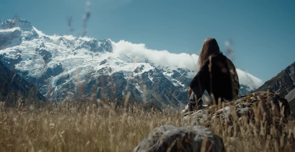 Woman sits on boulder in field looking toward snow covered mountain
